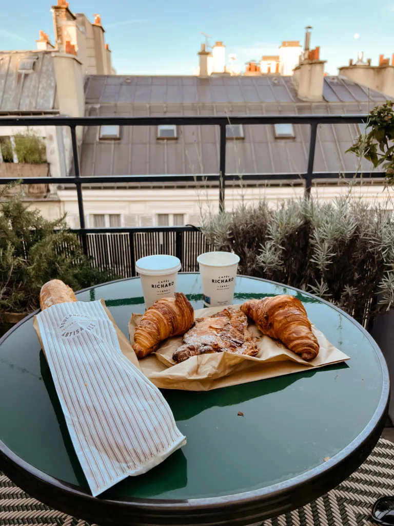 Baguette, two coffee cups, and pasteries sitting on a green table on a terrace.