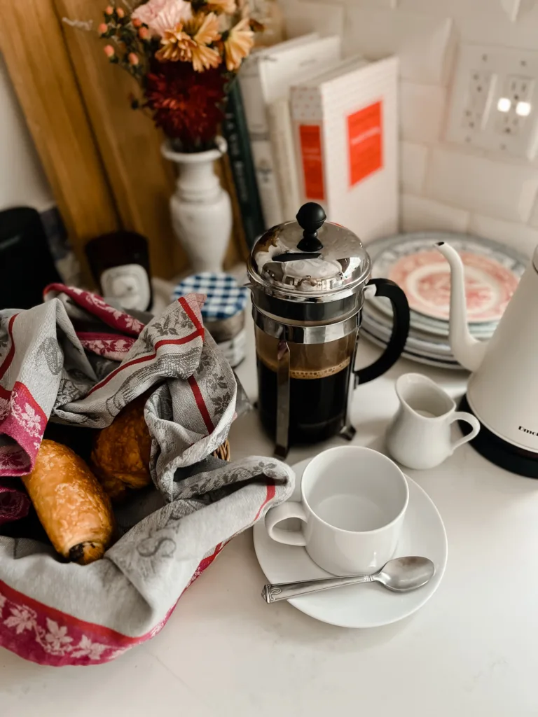 French press sitting on a counter with white coffee cup and croissants.
