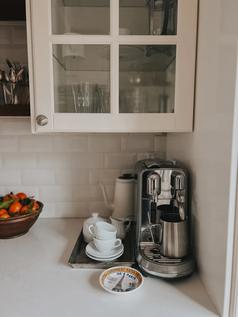 Nespresso machine on a kitchen counter with white espresso cups and bowl of oranges.