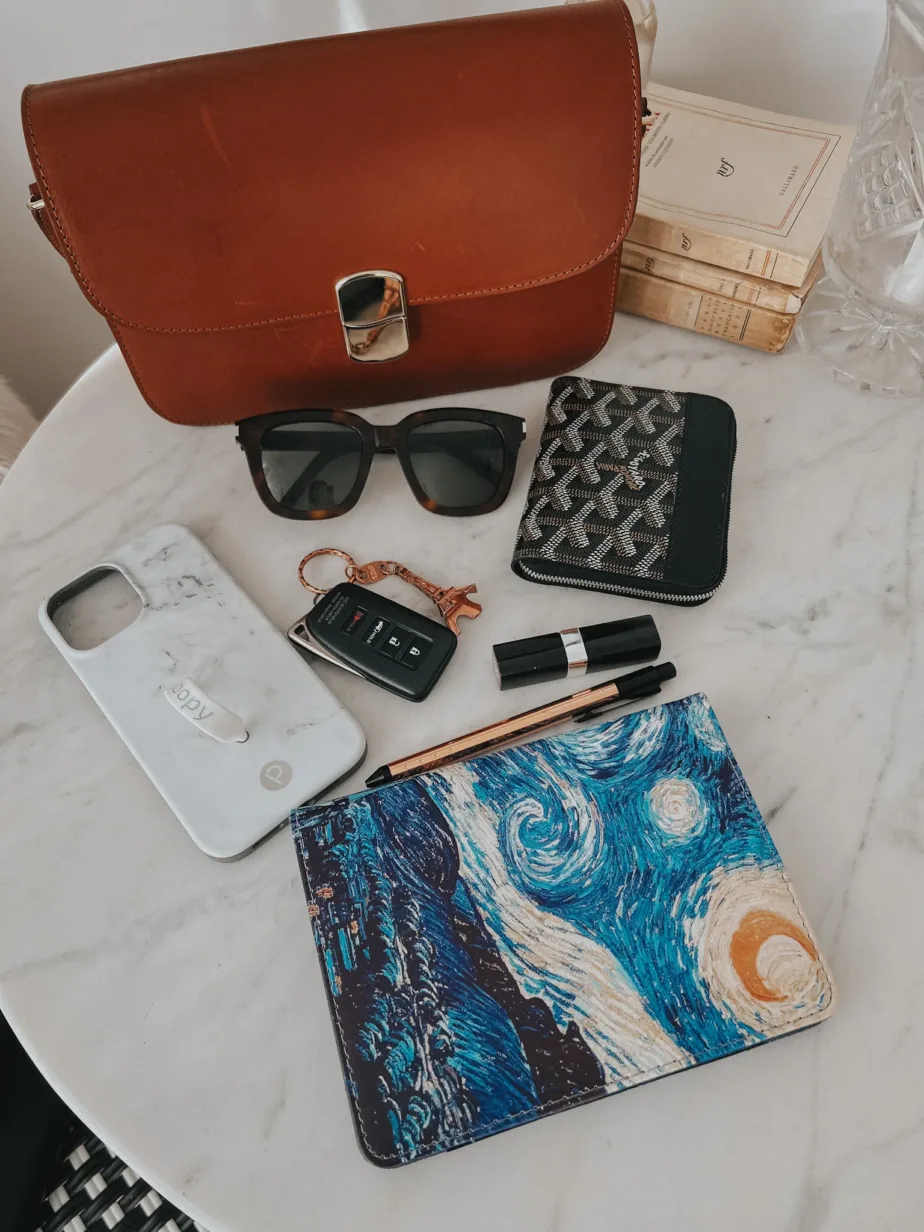 Brown leather bag sitting on table with sunglasses, wallet, lipstick, pen, kindle and phone cover.