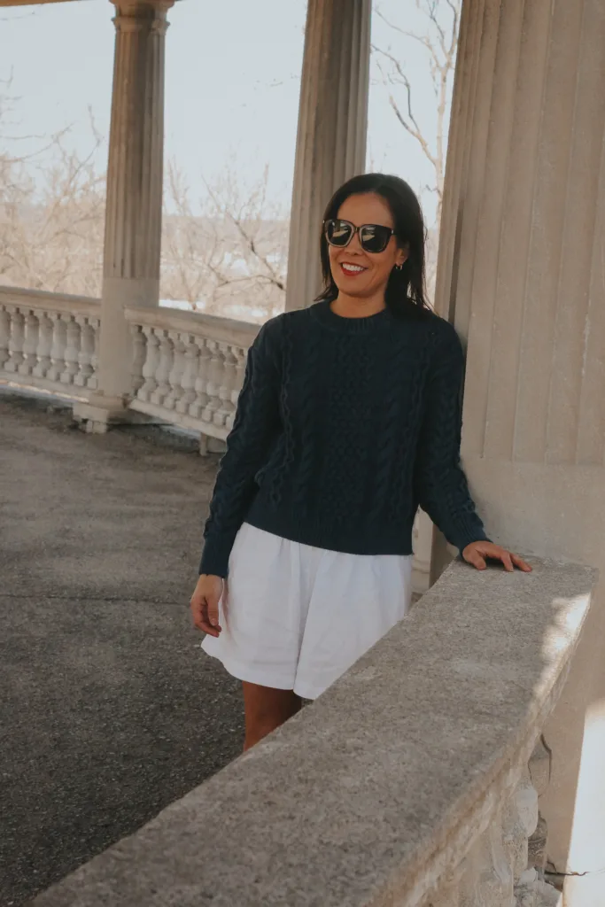 woman wearing navy sweater and white shorts leaning up against a wall.