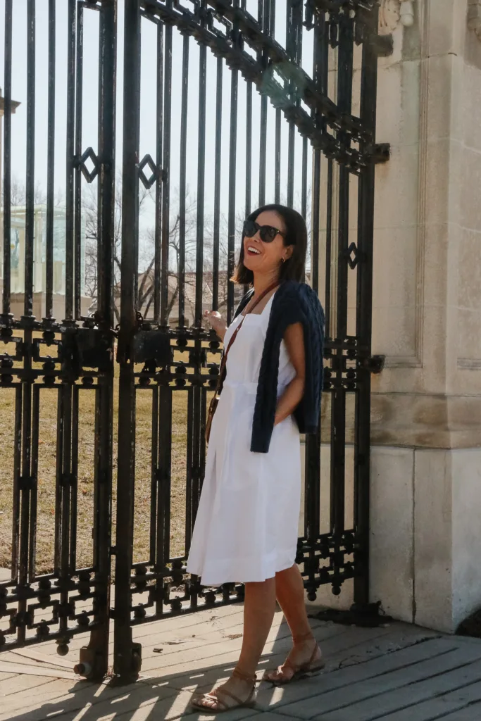 Woman wearing white linen sundress with navy sweater leaning against a black metal gate.