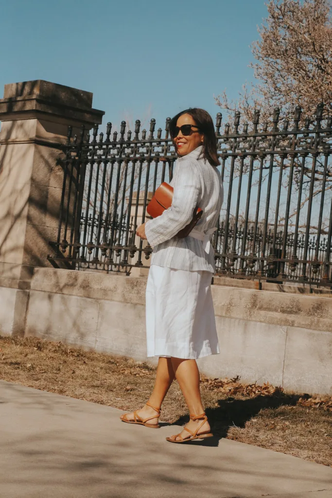 Woman wearing a blazer with white dress, sandals, and carrying a brown bag.