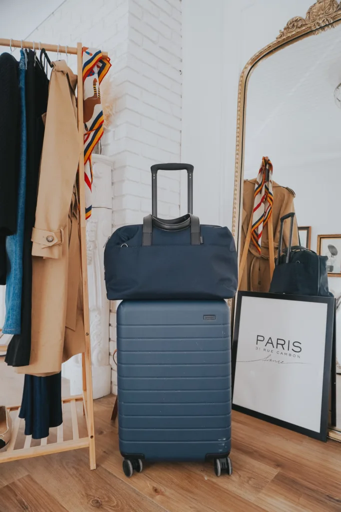 Blue suitcase and bag against a mirror with clothing rack in the back showing how to pack for paris in a carry-on.