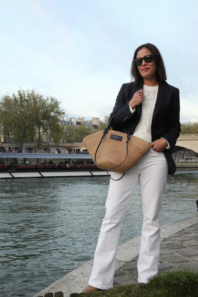 Woman wearing white denim, navy blazer, and straw tote along Seine.