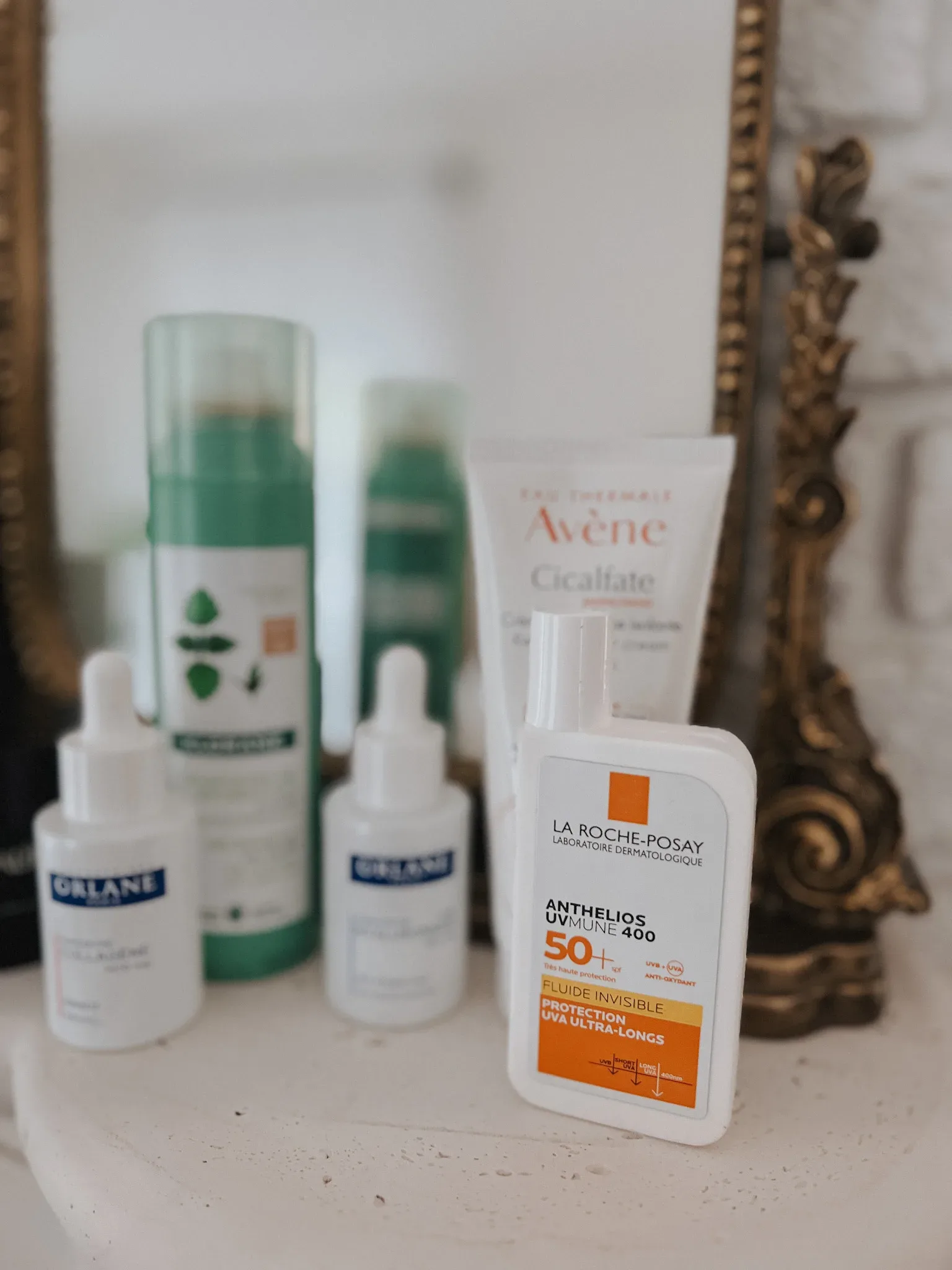 French beauty products in front of a gold mirror on a mantle. Sunscreen, dry shampoo.