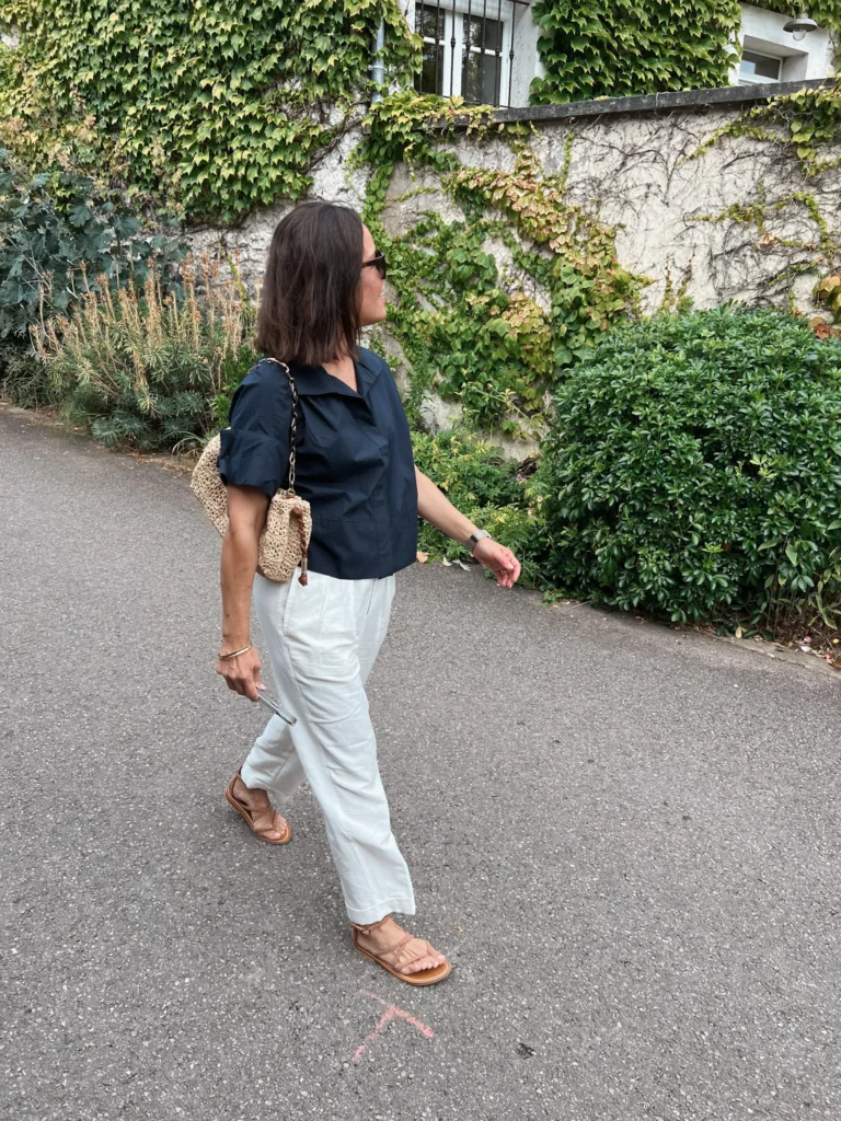 walking in Paris Woman with navy top and cream pants and sandals walking in front of green shrubs.