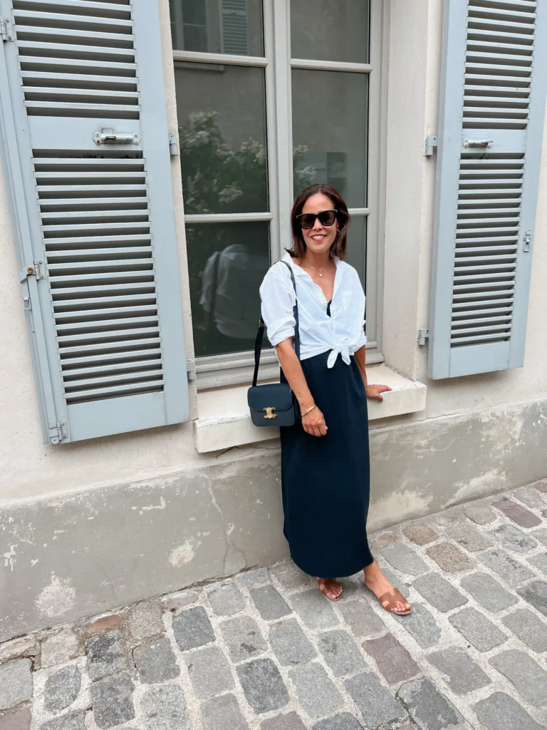 Woman wearing navy dress with white button down tied at waist standing against a window.