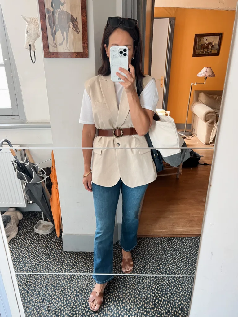 Woman wearing tan long blazer vest with white t-shirt taking a picture in the mirror.
