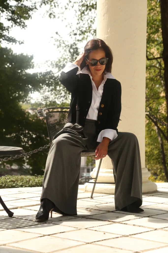 Woman sitting down in chair wearing grey pants, black cardigan, has hand in her hair.