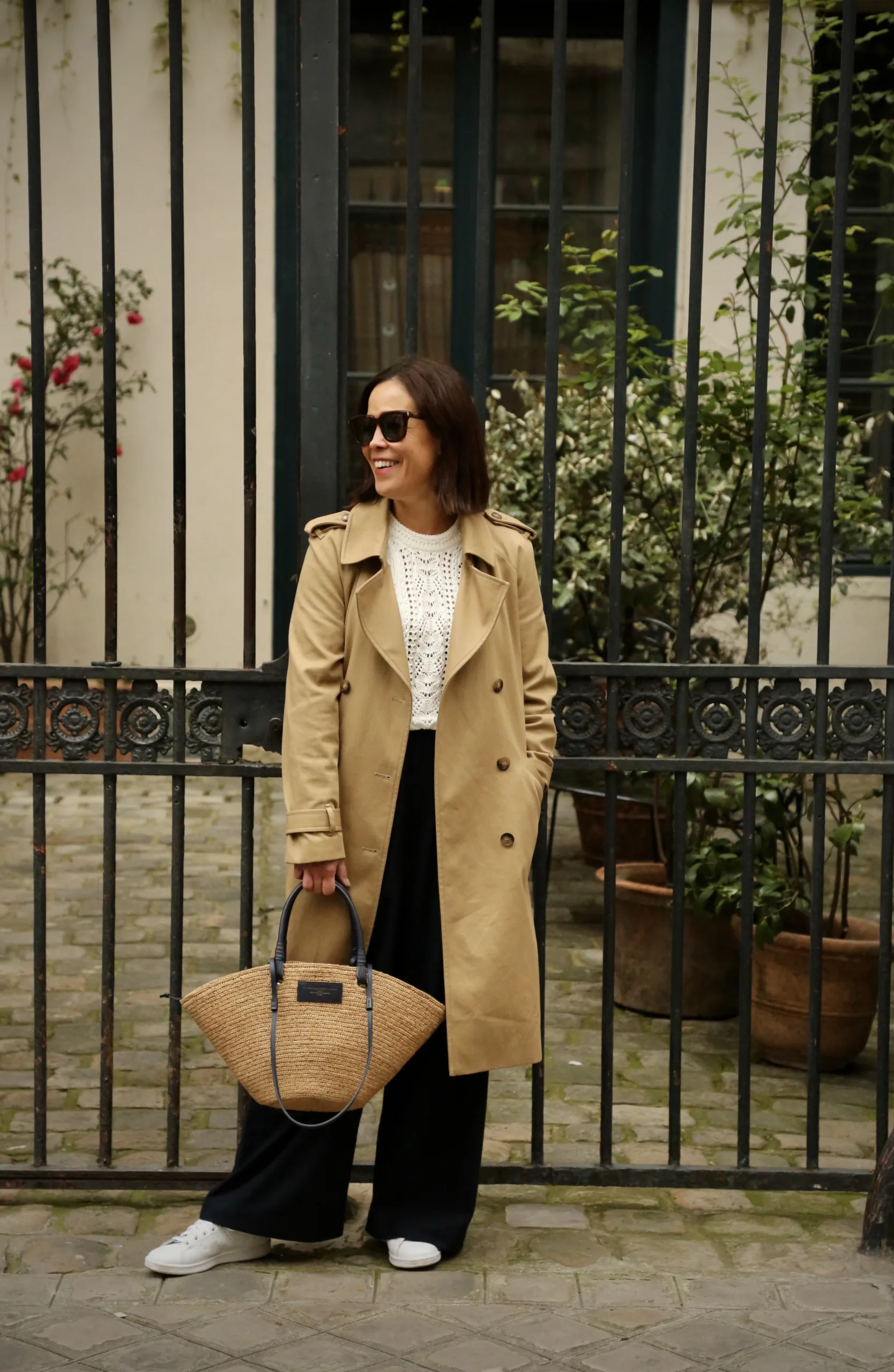 Woman wearing trench coat with sneakers standing against a fence.