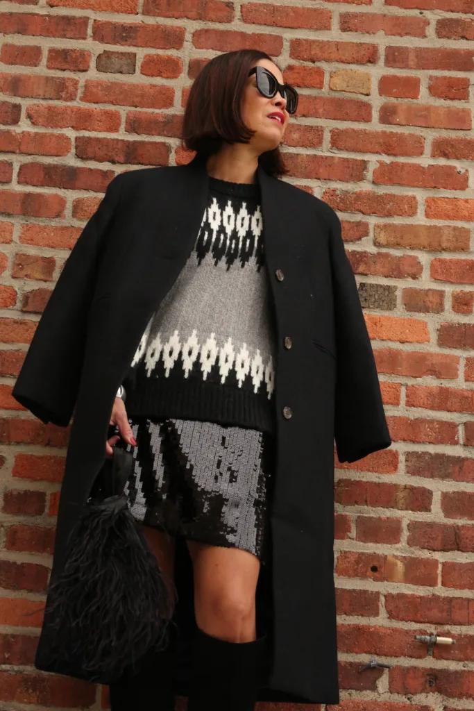 Woman in black coat and sweater standing against a wall with tall boots.