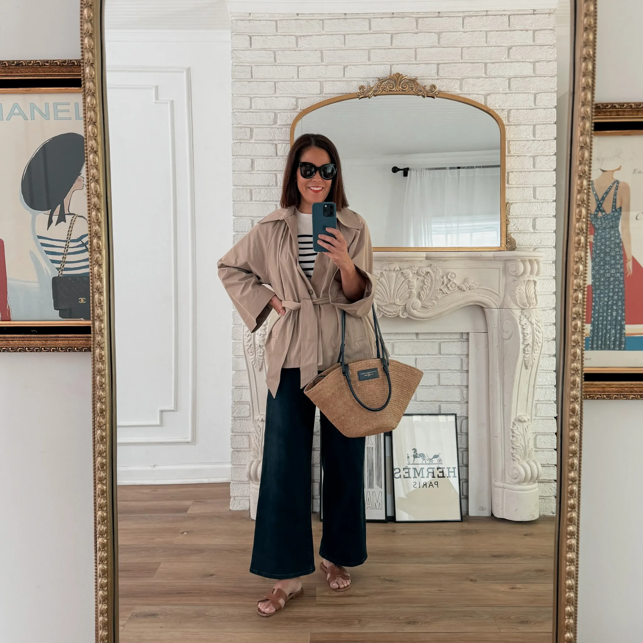 Woman wearing dark denim with sandals, and a short trench coat in front of a french mantle showing wardrobe basics for spring.