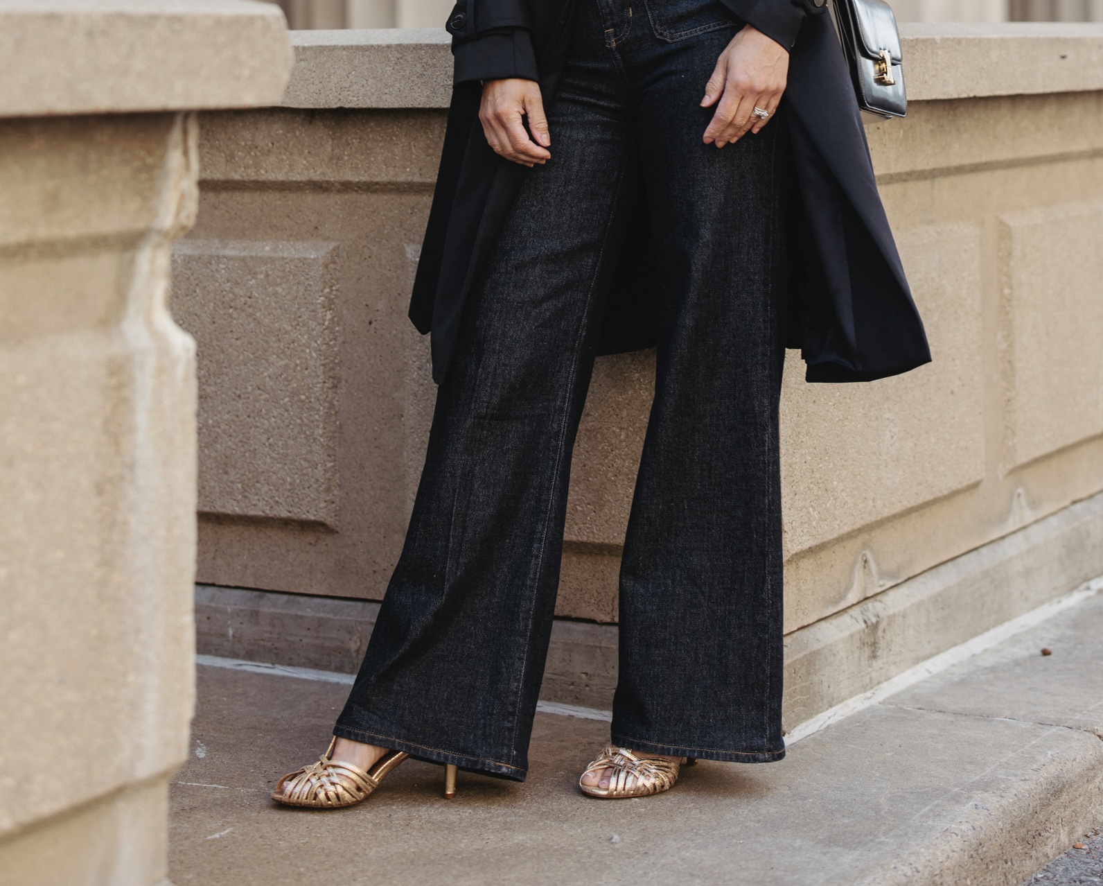 Woman wearing gold sandals standing against a wall.