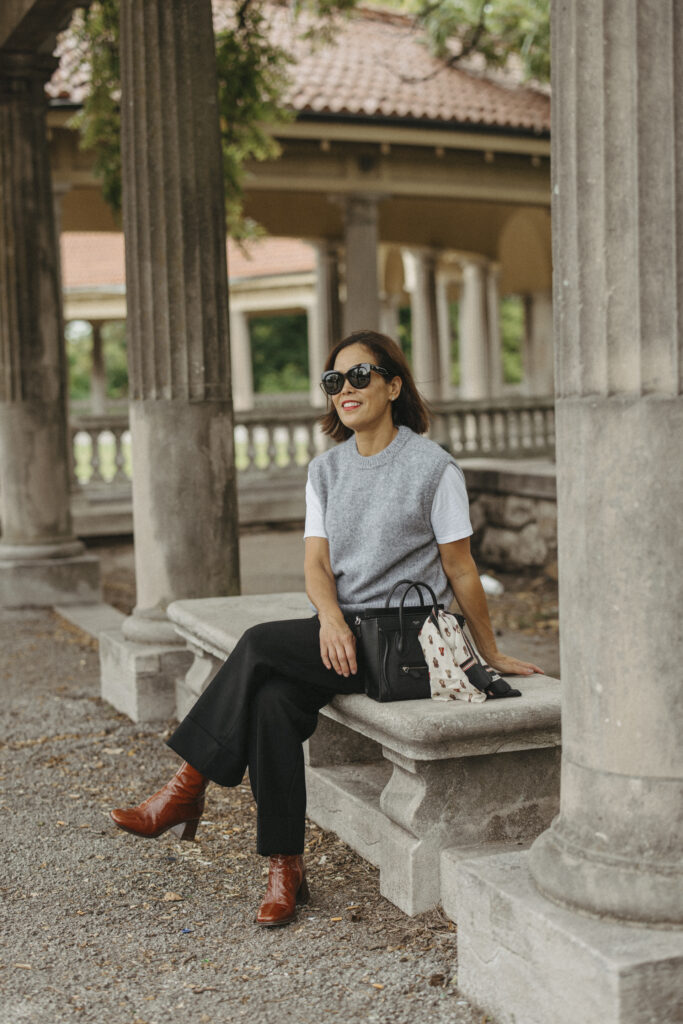 Lady wearing a grey vest with dark black pants sitting on a bench.
