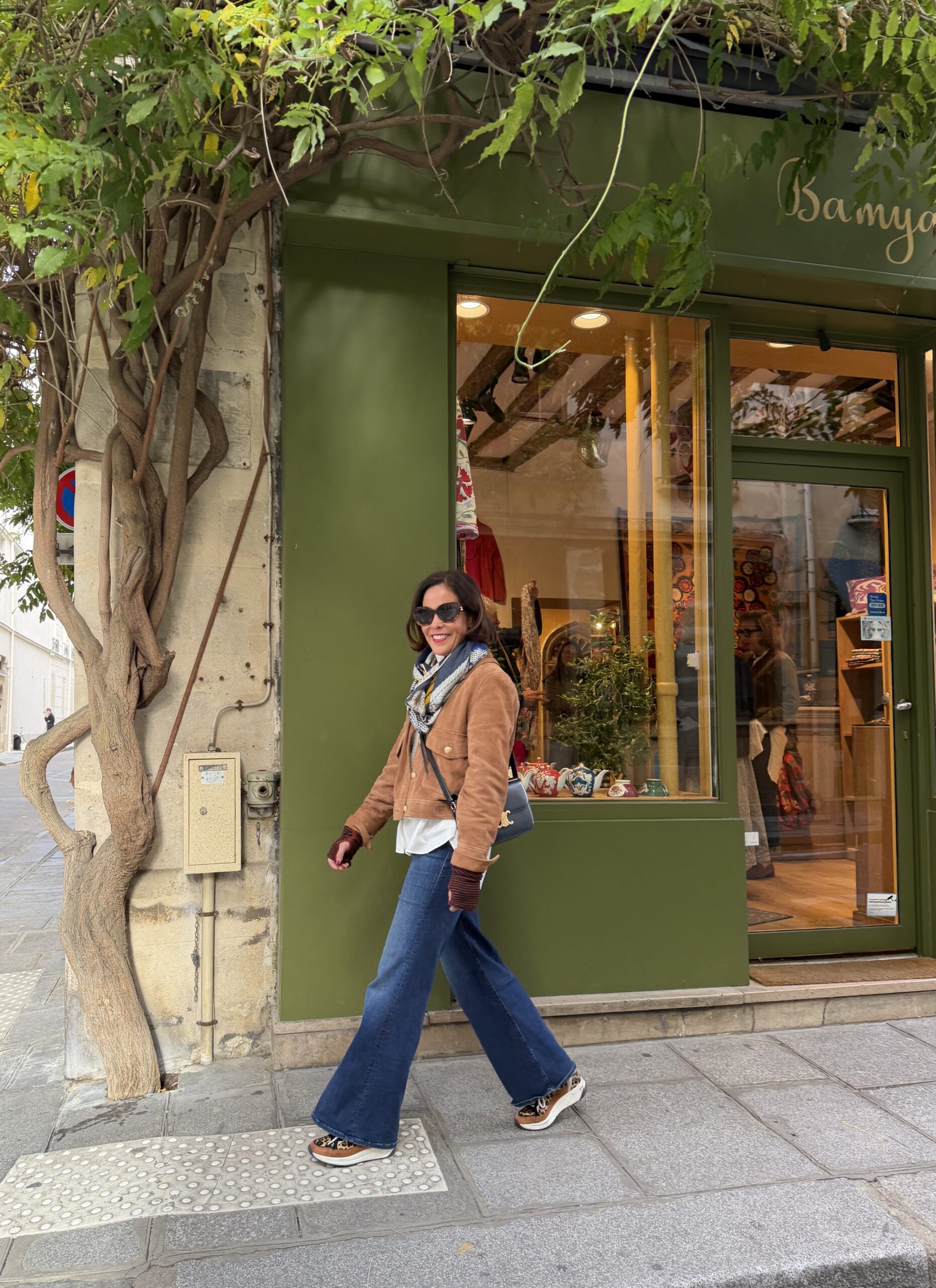 Woman wearing suede jacket with wide leg denim, sneakers, brown suede jacket and crossbody bag walking in front of a green wall.