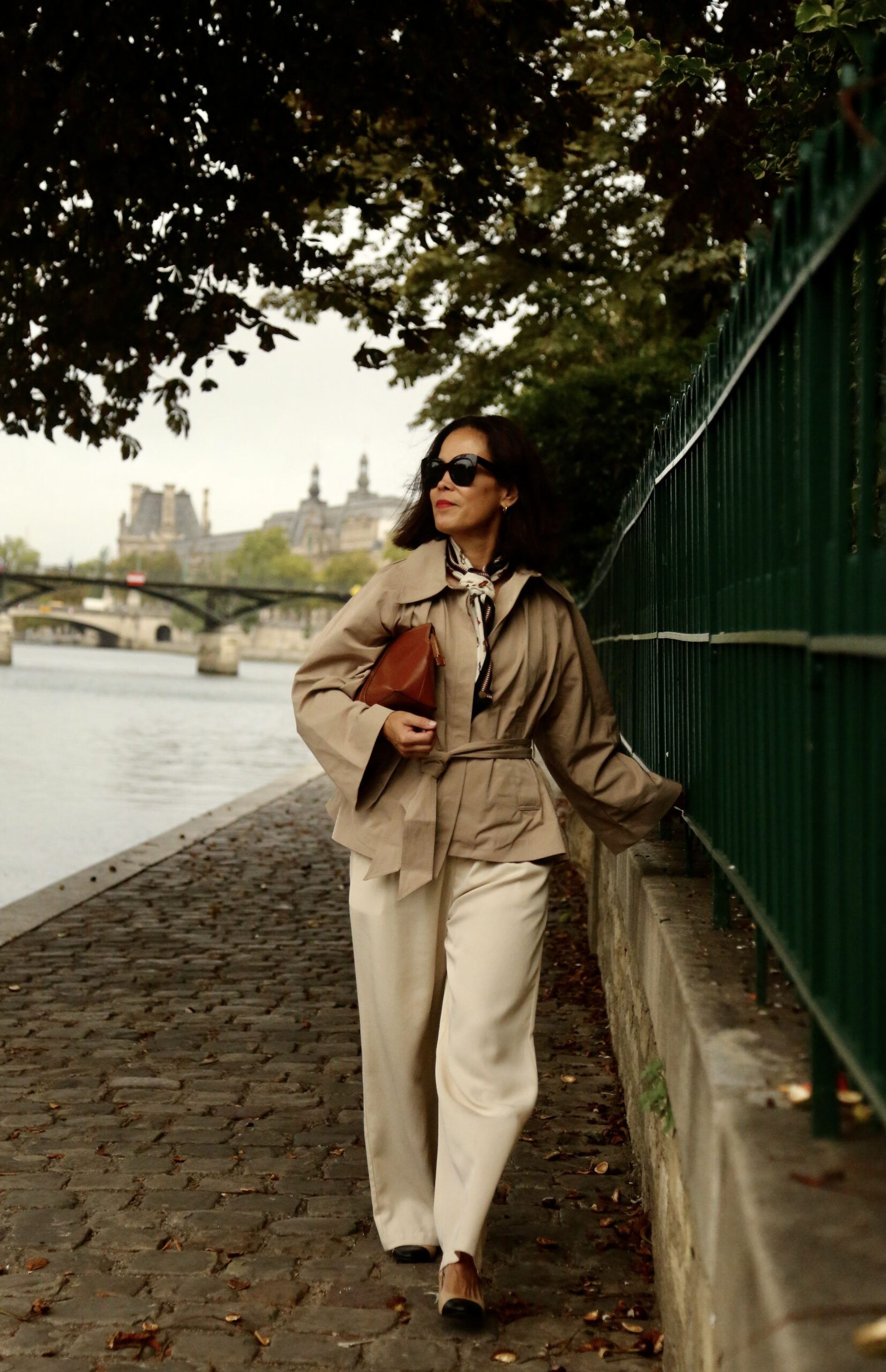 Woman walking along Seine in paris in a Paris fall outfit of trench coat and trousers with slingbacks.
