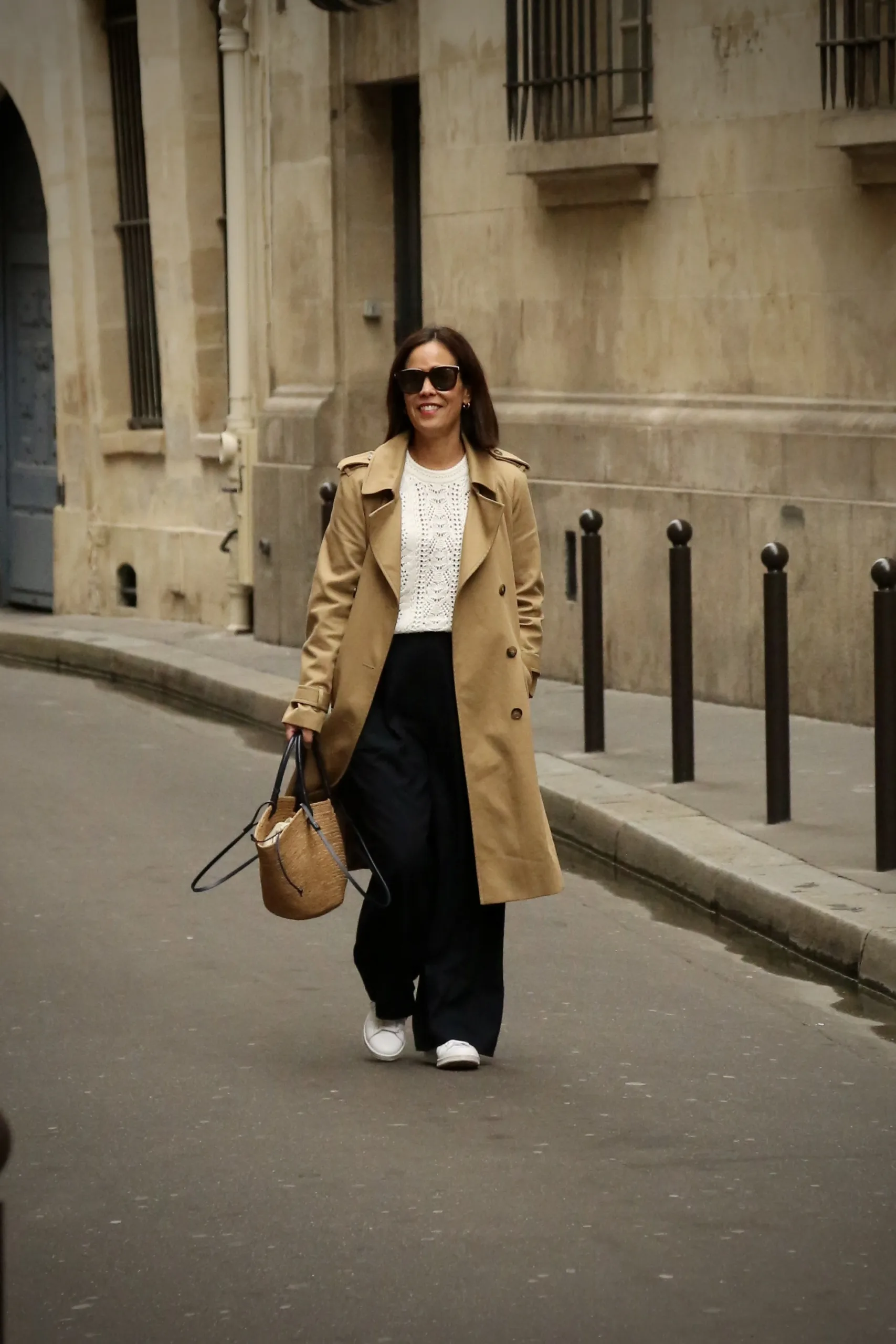 Woman walking down the street with a trench coat, pants, and sneakers.