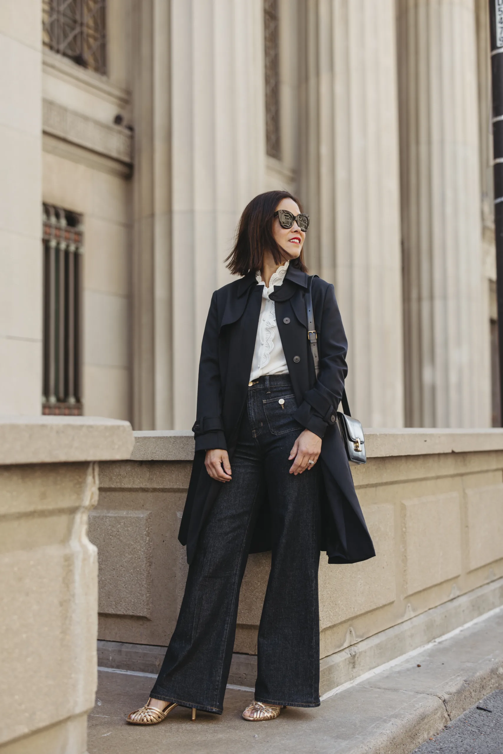 Woman wearing navy trench coat with dark denim, ivory top and gold sandals. 