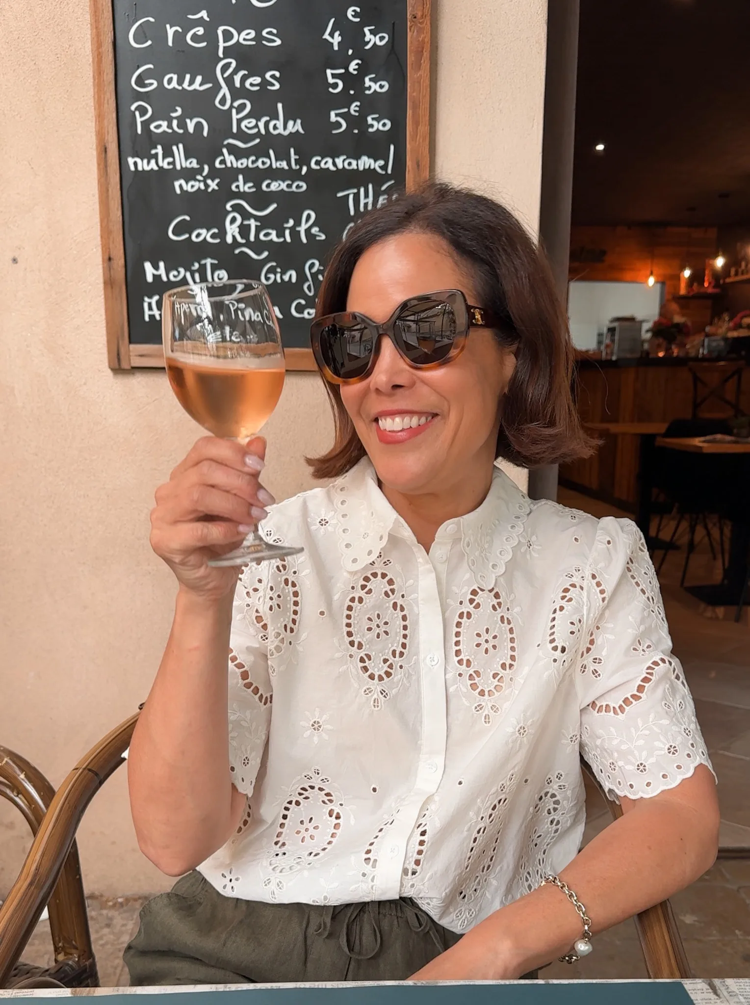 Woman wearing white eyelet top with linen pants and holding glass of wine.