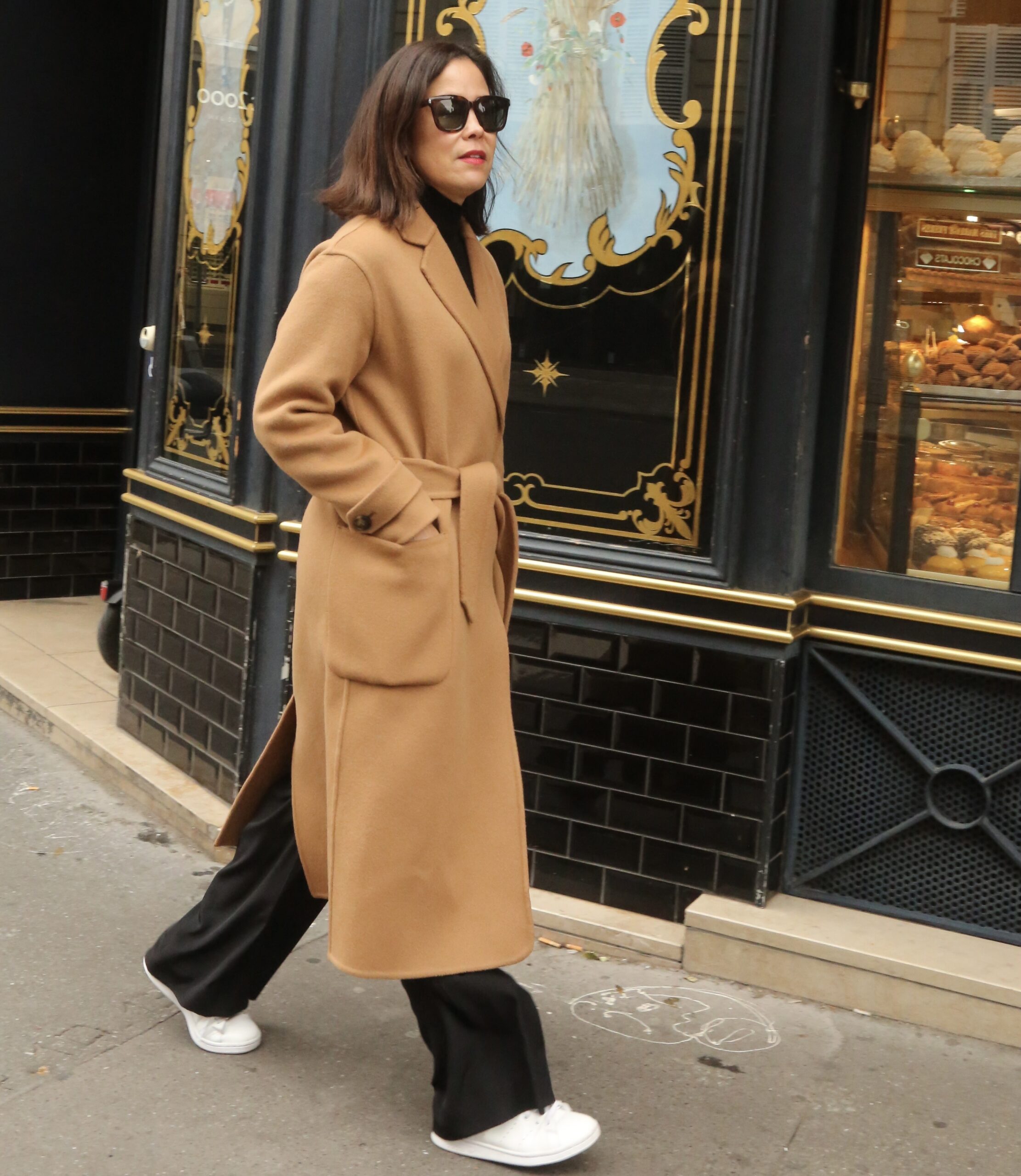 Woman with camel coat and white sneakers walking in Paris in January.