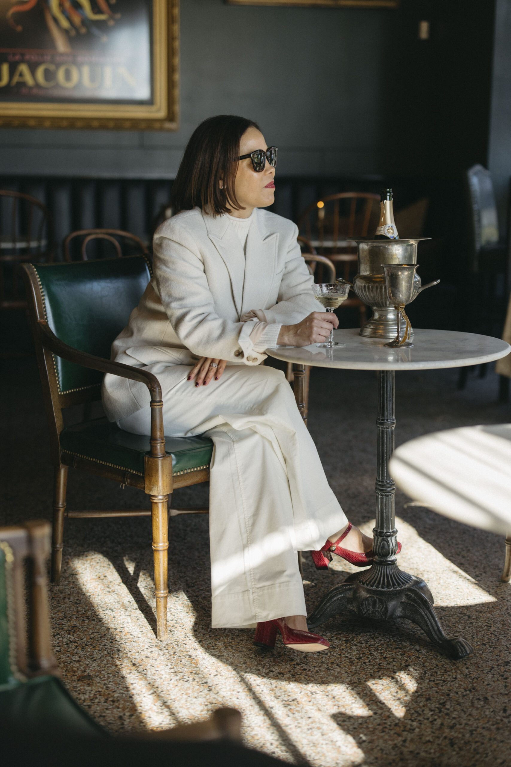 Woman wearing cream blazer with pants sitting in a champagne bar showing chic french girl party outfit.