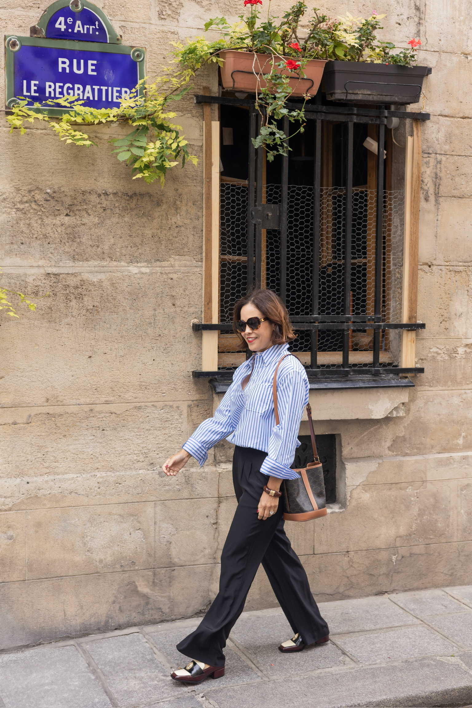 Woman wearing a striped button-up shirt, black trousers, and loafers while walking confidently in Paris